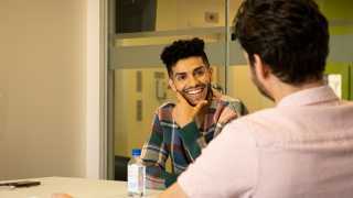 Mena Massoud | Aladdin actor Mena Massoud smiling and sitting at a table