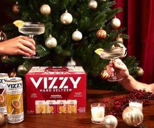 A Vizzy Mimosa Hard Seltzer case, two glasses cheers-ing in front of a Christmas tree